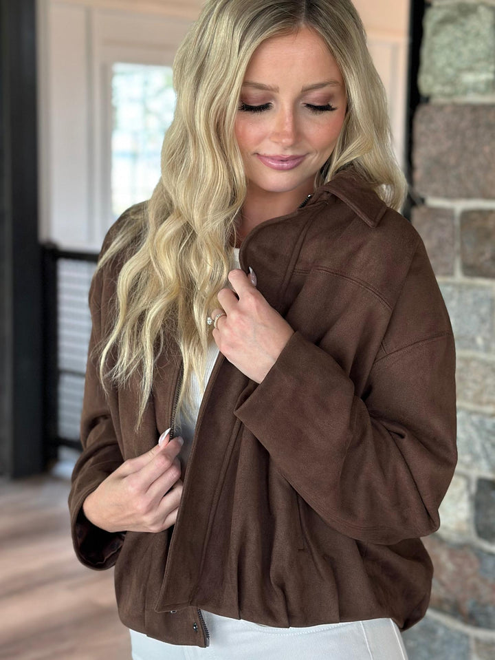 A blonde woman wearing a brown suede-like cropped jacket over a white top, standing indoors against a stone wall and window background.