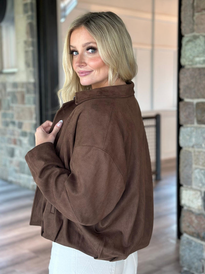 A woman with blonde hair wearing a brown cropped suede jacket and white pants, posing in a stone-walled indoor setting.