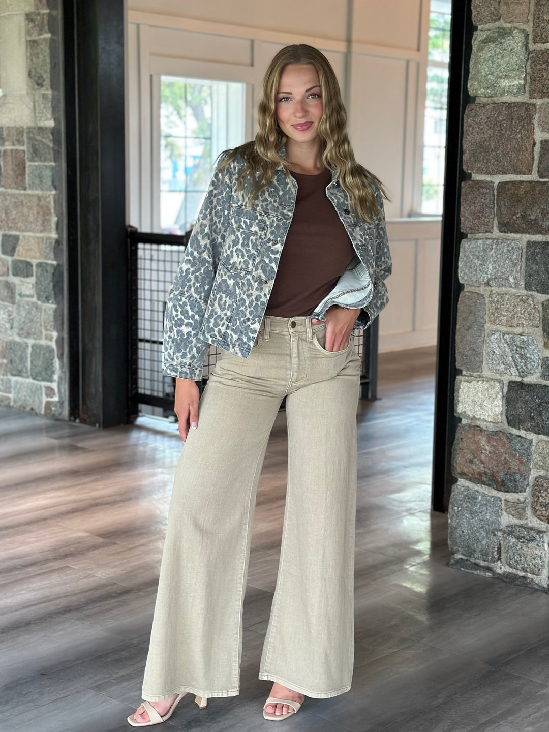 A woman modeling a light gray and white leopard print jacket over a brown top and beige wide-leg jeans, standing in a home interior with stone walls and wood floors.