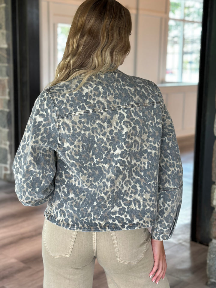A woman viewed from behind wearing a cropped denim jacket with a blue-gray and beige animal print, paired with beige pants, standing in an indoor setting.