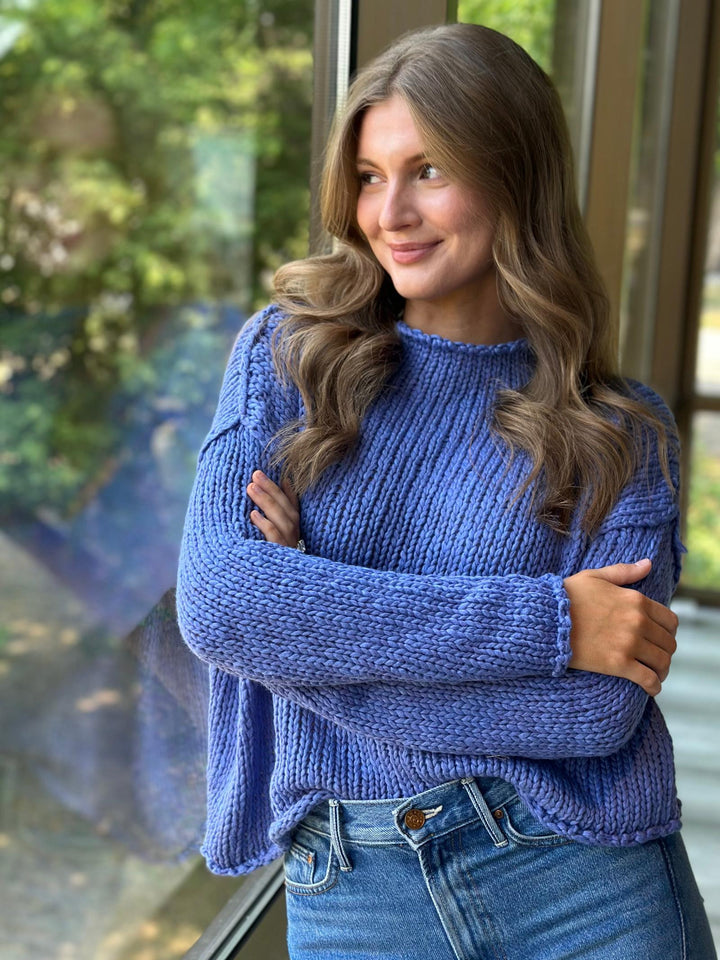 A woman with long wavy brown hair wearing a chunky blue ribbed knit sweater and blue jeans, standing with arms crossed in front of a window with a blurred outdoor background.