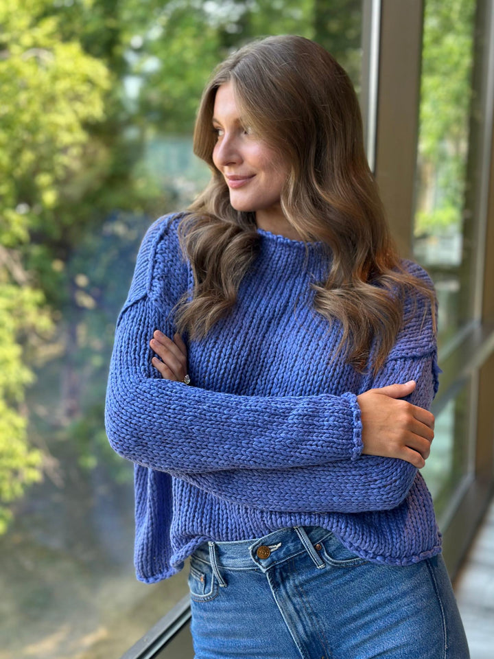 A woman wearing a chunky blue knit sweater and blue jeans, standing with arms crossed in front of a window with a blurred green outdoor background.