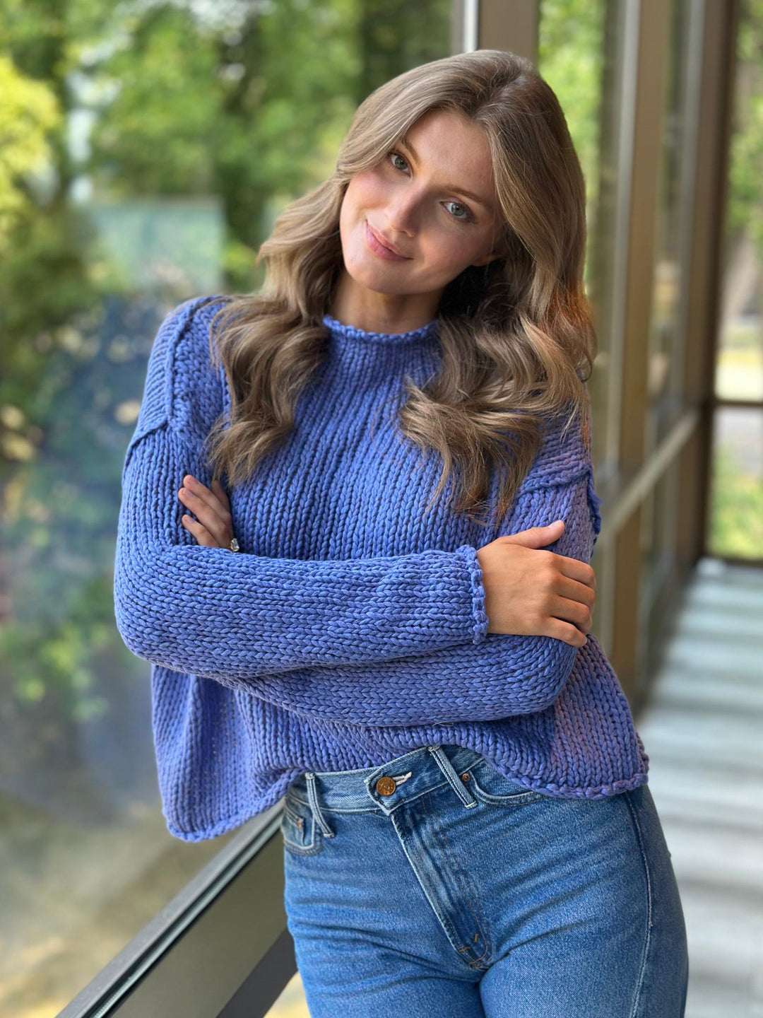 A woman with long wavy brown hair wearing a textured blue knit sweater and blue jeans, posing with her arms crossed in front of a large window with green foliage visible outside.