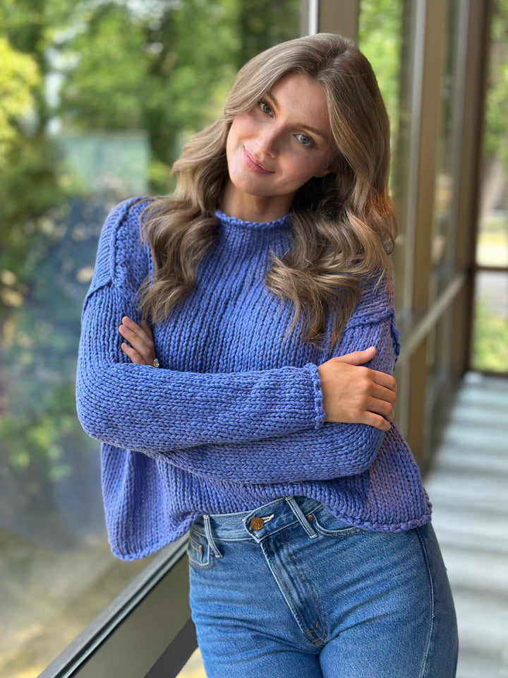 A woman with long wavy brown hair wearing a textured blue knit sweater and blue jeans, posing with her arms crossed in front of a large window with green foliage visible outside.