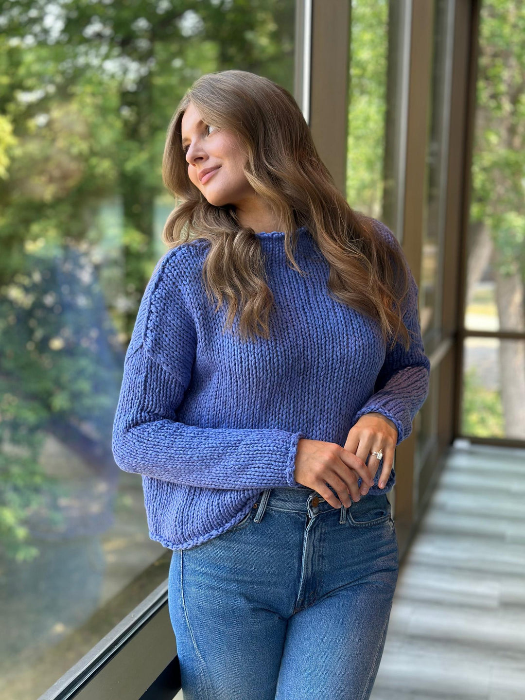A woman wearing a chunky blue cable-knit sweater and blue jeans, standing on a covered walkway with large windows and green trees visible outside.