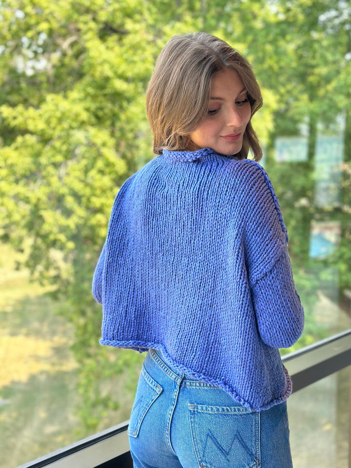 A woman wearing a blue chunky knit cropped sweater and light blue jeans, photographed from behind in an outdoor setting with green trees in the background.