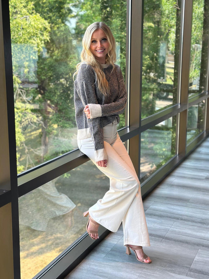 A woman posing in a modern indoor setting with large windows, wearing a gray knit sweater, cream wide-leg pants, and nude high-heeled sandals.