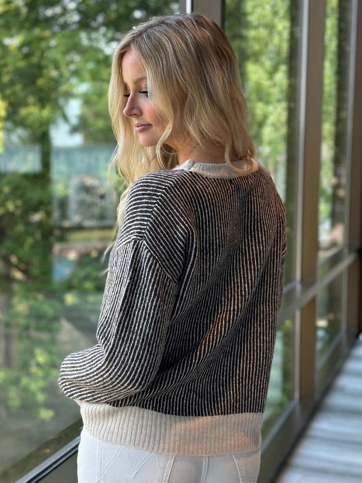 A woman with blonde hair is shown from the back wearing a black and white vertically striped knit sweater with a cream ribbed hem, standing by a large window with green trees visible outside.