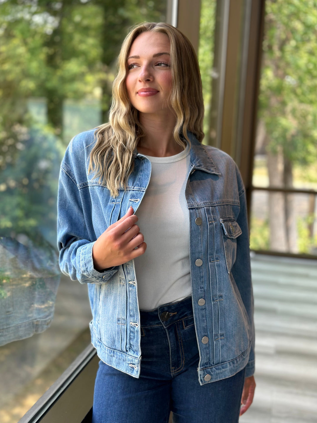 A woman wearing a light-wash blue denim jacket over a white t-shirt and dark blue jeans, standing in front of a large window with a blurred outdoor background.