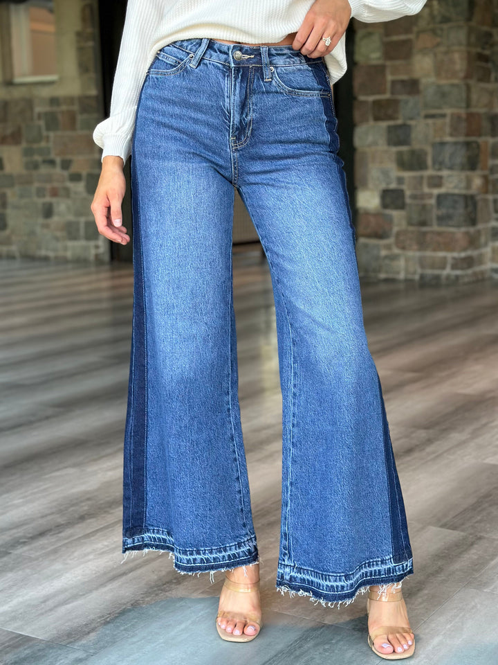 A person wearing medium-wash blue flare jeans with a frayed hem, paired with a white long-sleeve top and clear high-heel sandals, standing on a wood floor in front of a stone wall.