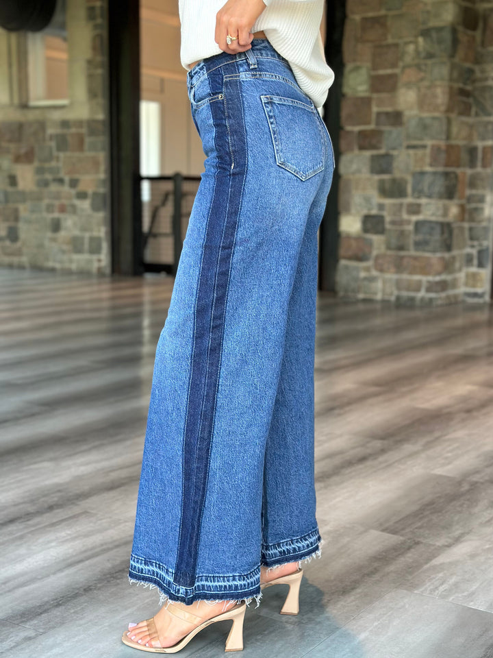 A person wearing high-waisted, wide-leg blue denim jeans with a double-stripe design down the side and frayed hems, paired with beige heeled sandals in an indoor setting with a stone wall and wood floor.