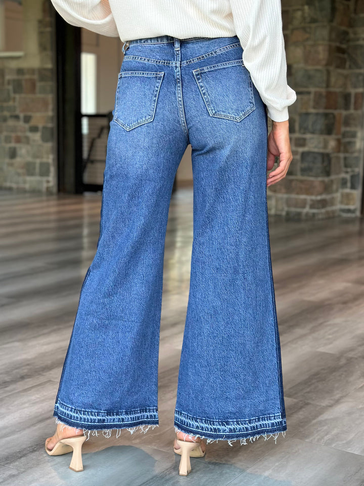 A person shown from the back wearing wide-leg blue denim jeans with a frayed hem, paired with a white long-sleeve top and beige heeled sandals in an indoor setting with a stone wall and wood floor.