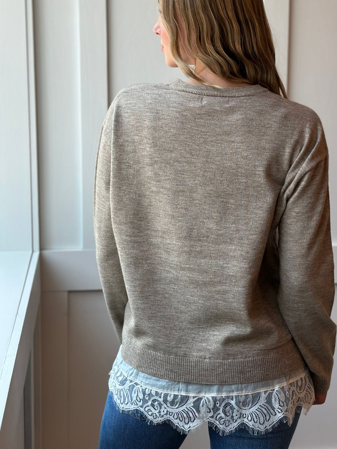 A woman shown from the back wearing a taupe heathered knit sweater over a light blue shirt with a white lace hem, paired with blue jeans.