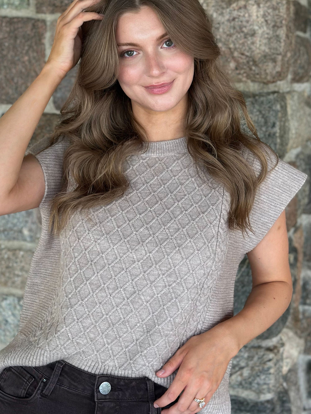 A woman with long wavy brown hair wearing a light beige textured knit top with cap sleeves, paired with dark jeans, standing against a rustic stone wall.