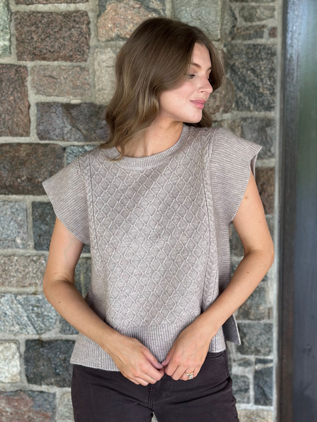 A woman wearing a light taupe knit top with textured diamond cable patterns and ruffled cap sleeves, standing against a rustic stone wall.