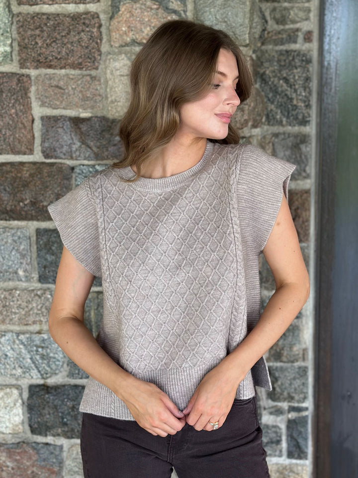 A woman wearing a light taupe knit top with textured diamond cable patterns and ruffled cap sleeves, standing against a rustic stone wall.