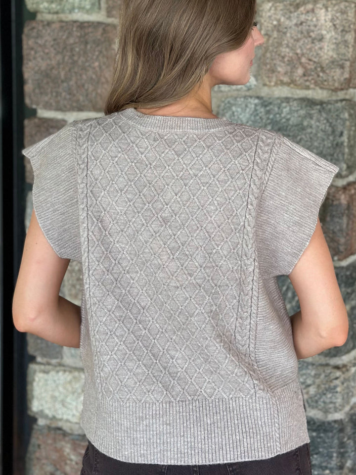 A woman viewed from the back wearing a light gray short-sleeve knit top with a textured diamond pattern on the back, standing against a stone wall.