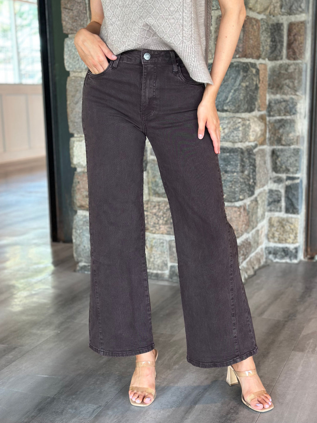 A person wearing dark charcoal wide-leg jeans with a gray textured top and clear-strap heeled sandals, standing in front of a stone wall and wood floor.
