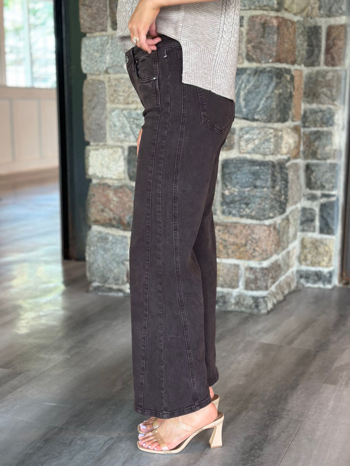 A person wearing dark charcoal wide-leg jeans with a light gray knit top and clear heeled sandals, standing in front of a stone pillar and wood floor.