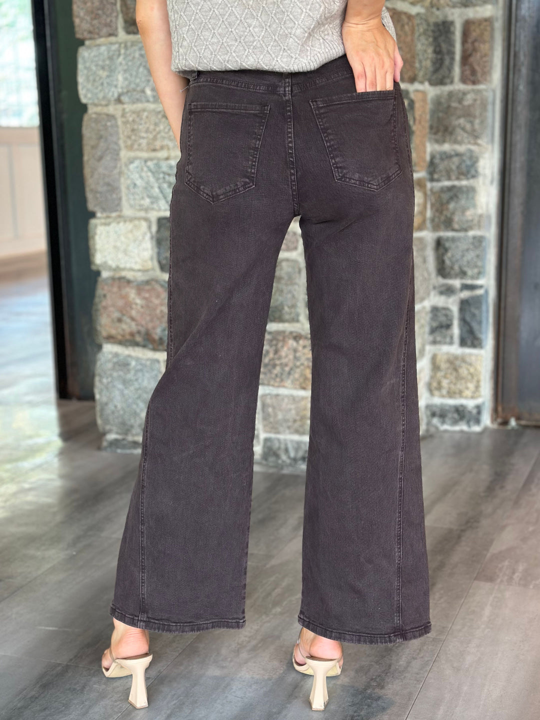 A person shown from the back wearing dark charcoal wide-leg jeans, a light gray textured top, and beige heeled sandals, standing in front of a stone wall.