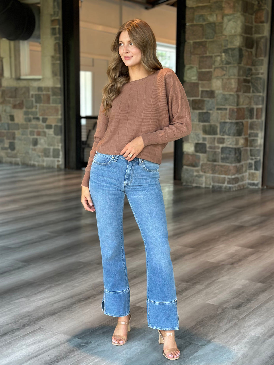 A woman modeling a brown long-sleeve knit top and light blue flare jeans with rolled cuffs, standing in a modern indoor space with stone walls and wood-look flooring.