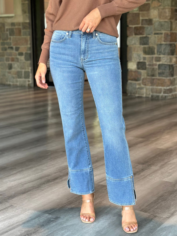 A person wearing light blue cropped flare jeans with rolled cuffs and side slits, paired with a brown long-sleeve top and clear high-heeled sandals, standing on a wood floor in front of a stone wall.