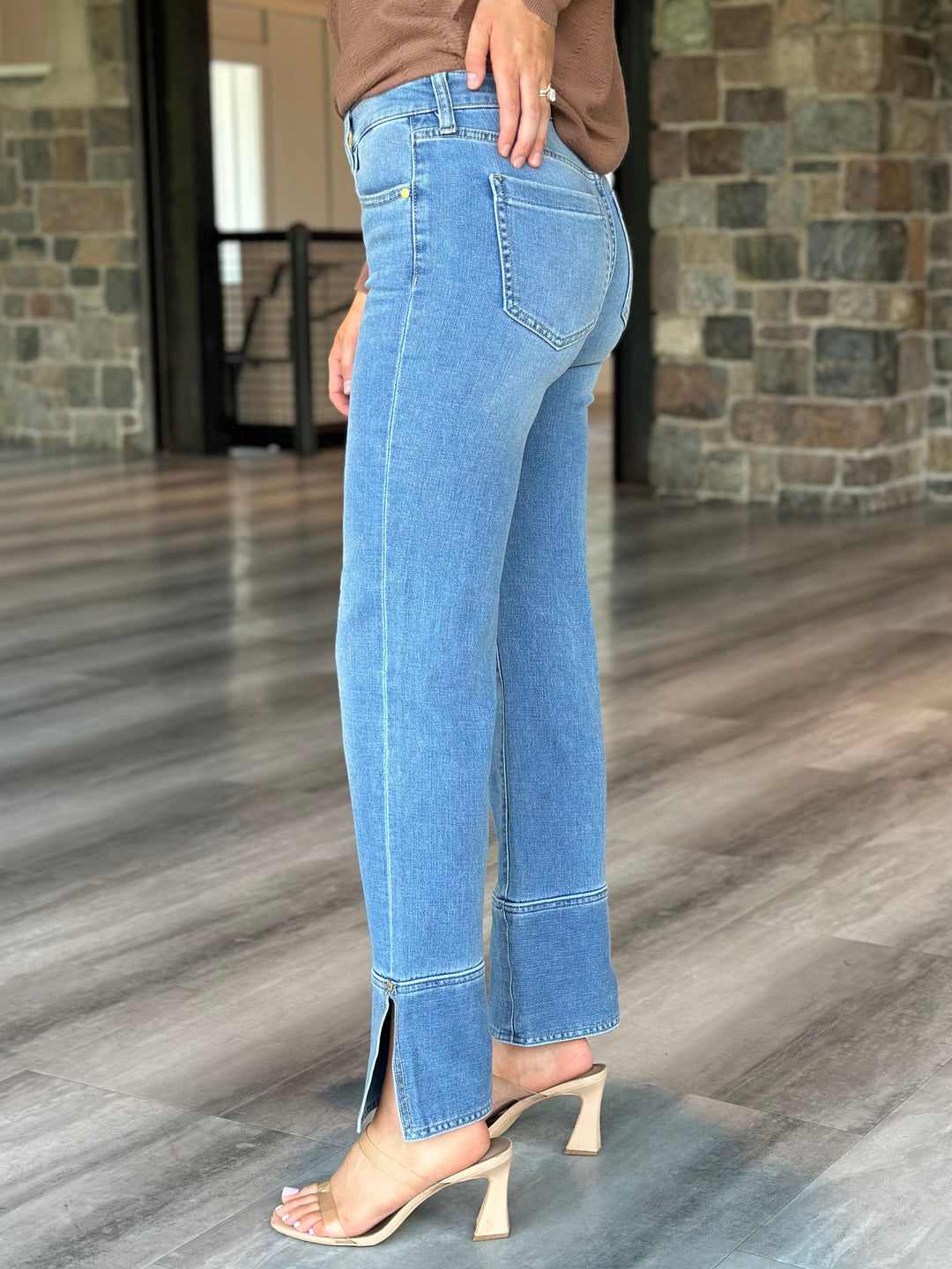 A person wearing light blue cropped jeans with side slits and a brown top, shown from the back in an indoor setting with wood flooring and stone walls.