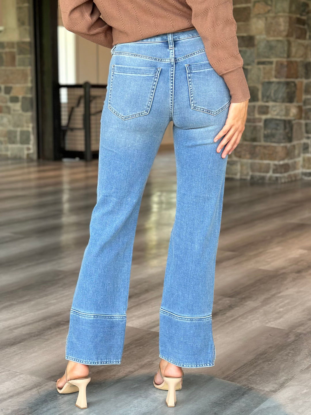 A person shown from the back wearing light blue wide-leg jeans with a cuffed hem, paired with a brown sweater and beige heeled sandals in an indoor setting with wood floors and a stone wall.
