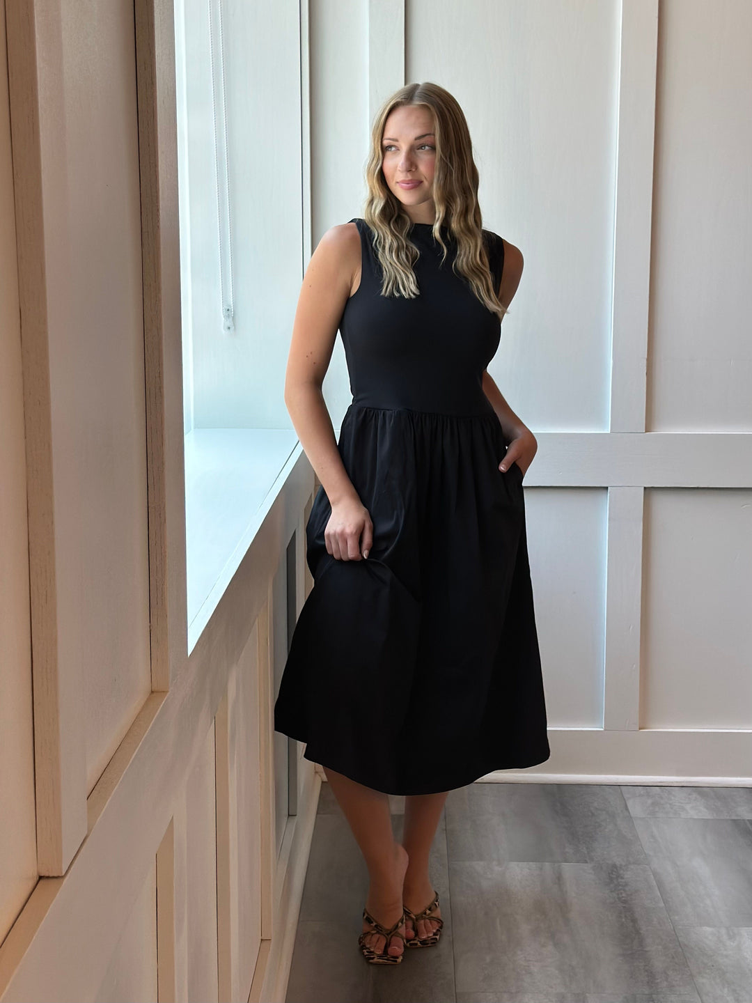 A woman modeling a sleeveless black midi dress with a high neckline and side pockets, standing in a brightly lit room with light wood paneling and gray flooring.