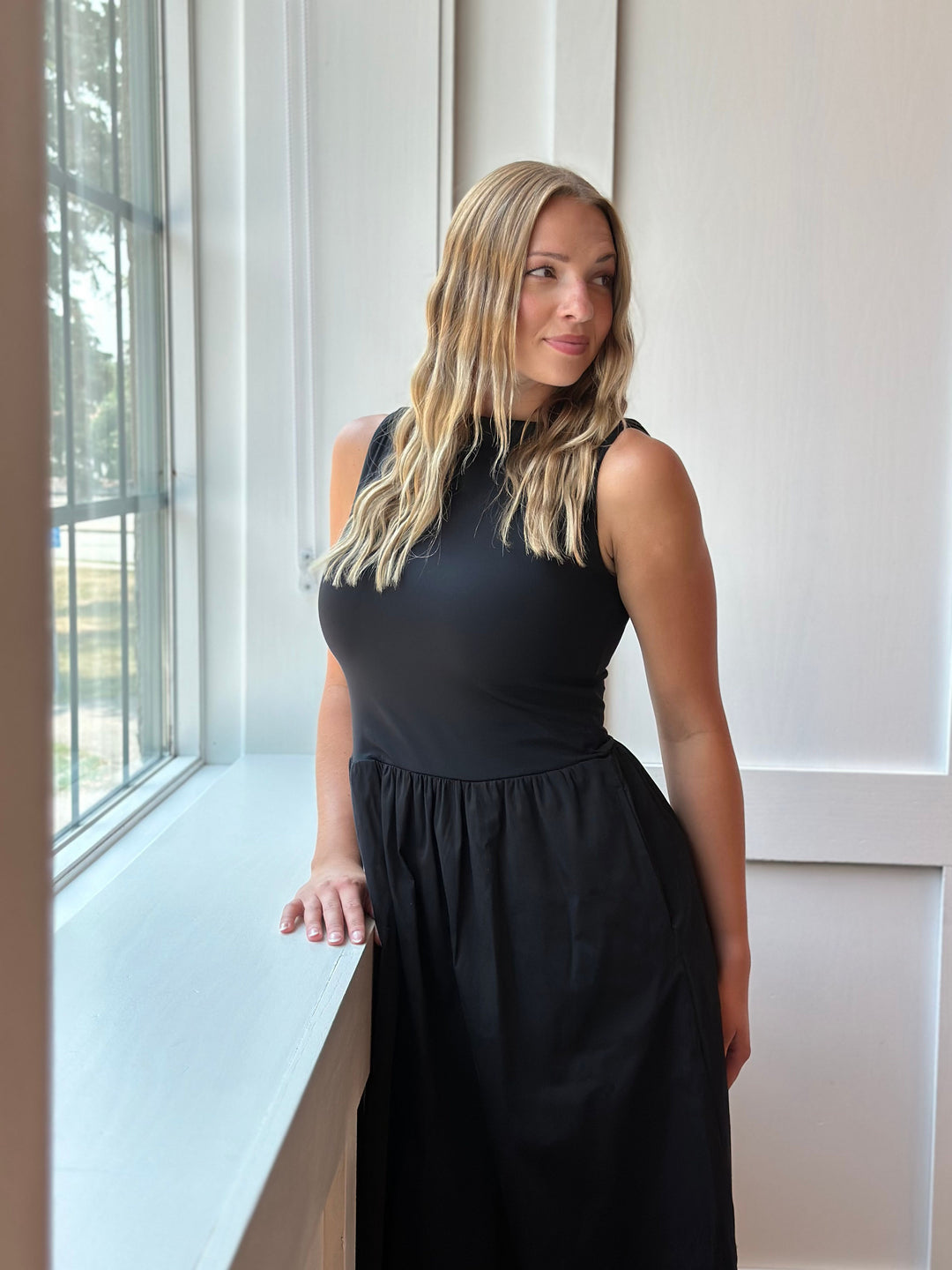 A woman with blonde hair wearing a sleeveless black midi dress, standing next to a window in a bright room.