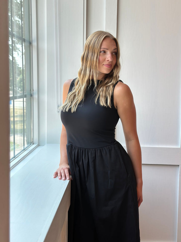 A woman with blonde hair wearing a sleeveless black midi dress, standing next to a window in a bright room.