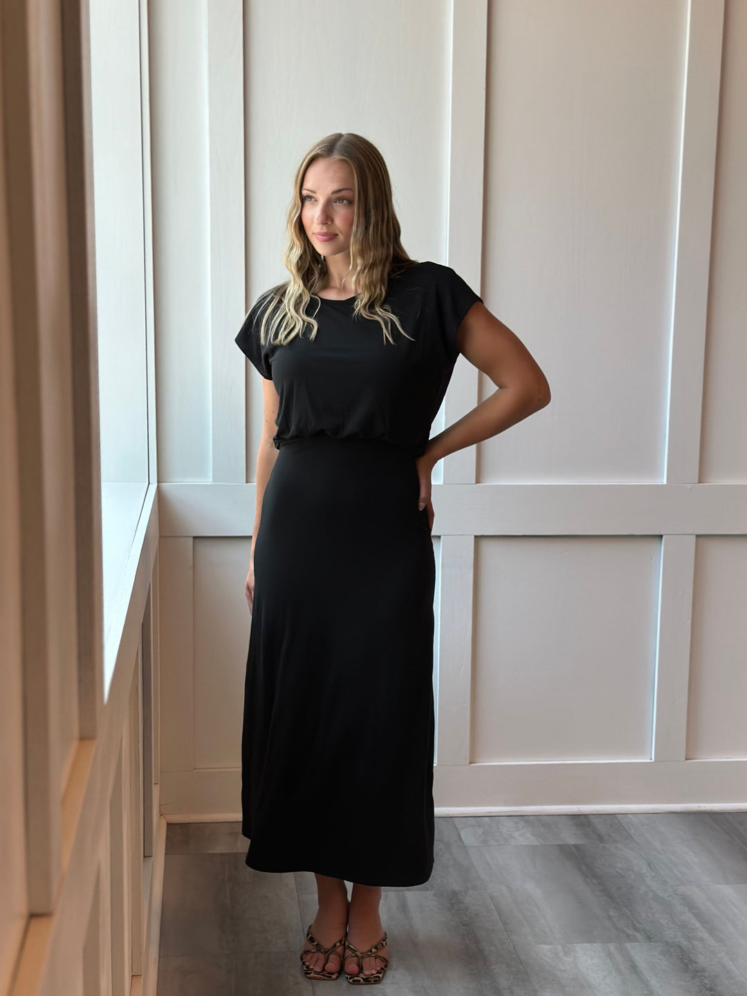 A woman modeling a black short-sleeve maxi dress with a gathered waist and side pockets, standing in a brightly lit room with white paneled walls and gray wood-look flooring.