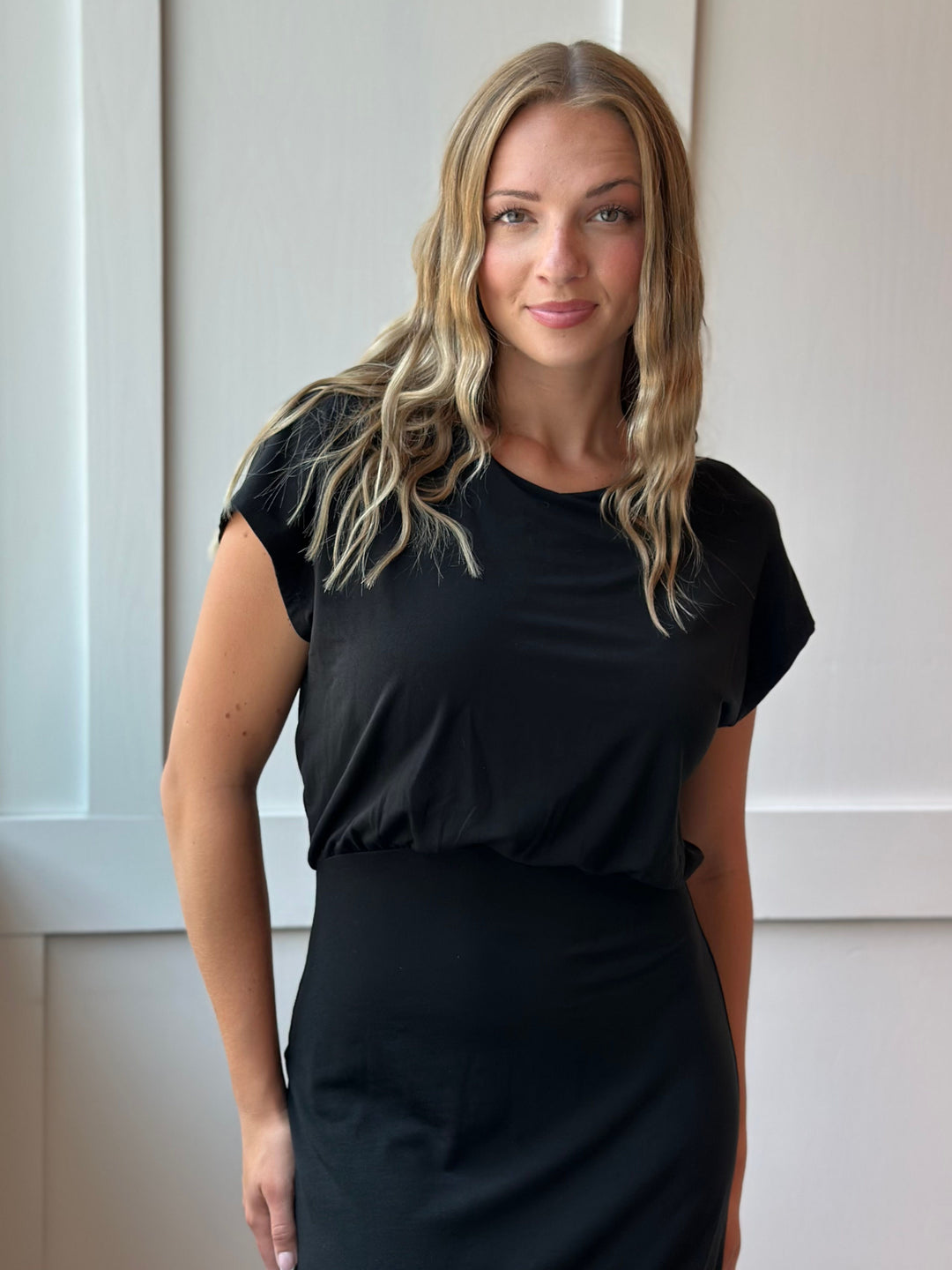 A woman with blonde hair wearing a black short-sleeve top and a black skirt, standing against a light-colored paneled wall.
