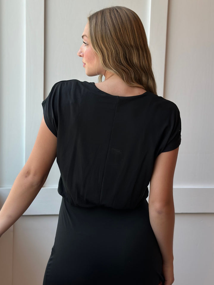 A woman shown from the back wearing a black short-sleeve top and black skirt, standing indoors against a light neutral paneled wall.