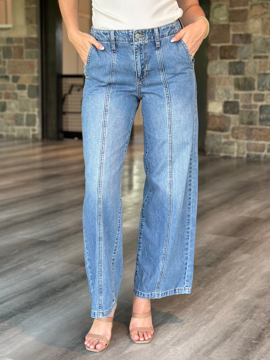 A person wearing light blue wide-leg denim jeans with a white top and clear-strap heeled sandals, standing on a wood-look floor in front of a stone wall.