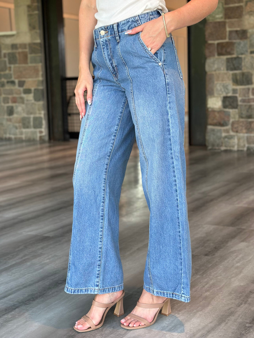 A person wearing light blue wide-leg denim jeans with a white top and beige heeled sandals, standing in an indoor space with stone walls and wood flooring.