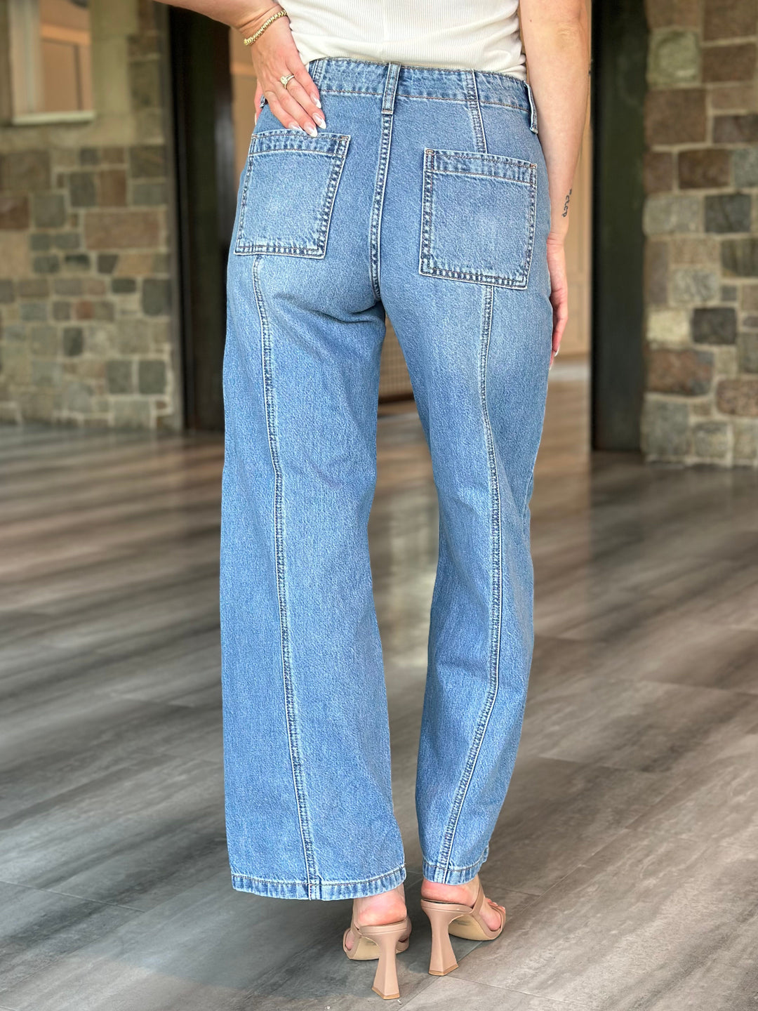A person shown from the back wearing light blue wide-leg denim jeans with a cream top and beige heeled sandals, standing on a wood-look floor in front of a stone wall.