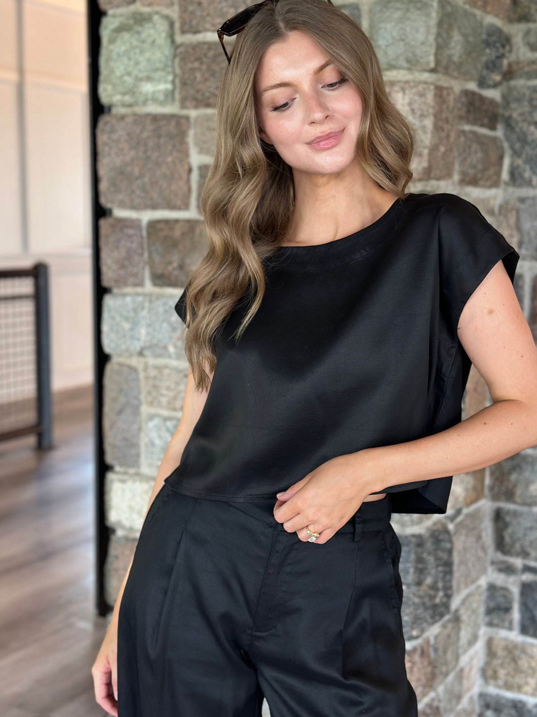 A woman wearing a black short-sleeve satin top and black trousers, standing in front of a stone wall.