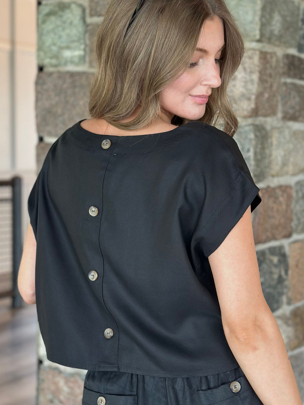 A woman wearing a black short-sleeve top with a back button closure, photographed from the back against a stone wall.