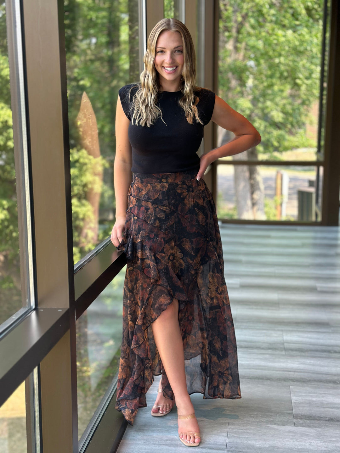 A woman stands barefoot on a tiled indoor porch wearing a black sleeveless top and a long, flowy high-low skirt with a dark floral pattern in brown and rust tones.