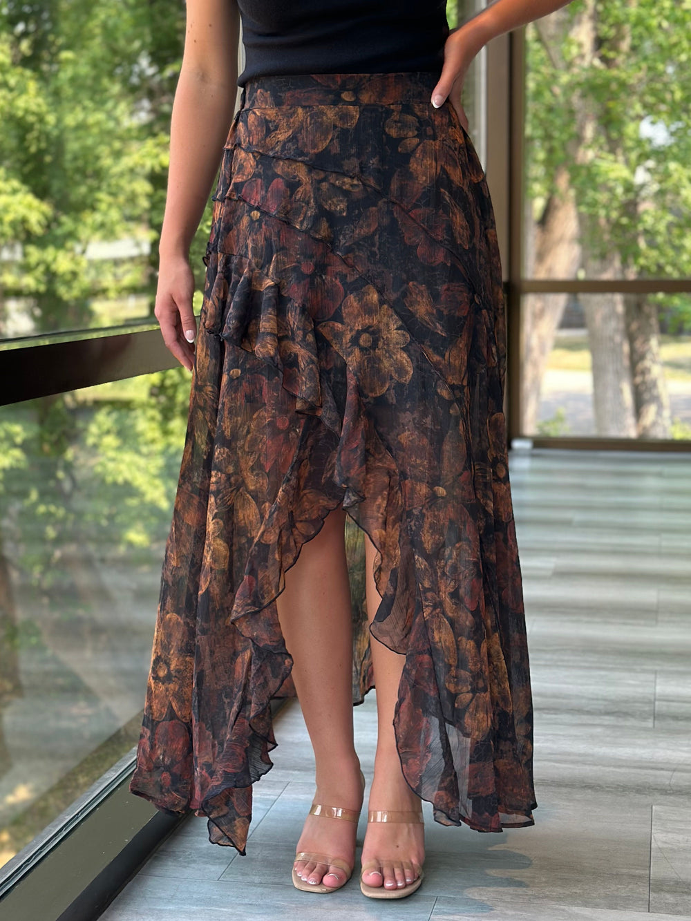 A woman wearing a long, ruffled high-low skirt with a dark floral pattern in brown, rust, and black, paired with clear-strap heels, standing on a tiled balcony with a green outdoor background.