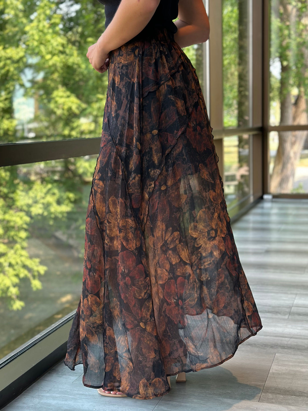 A woman wearing a long, flowy maxi skirt with an abstract floral pattern in dark brown, rust, and black, standing on a balcony with large windows overlooking green trees.