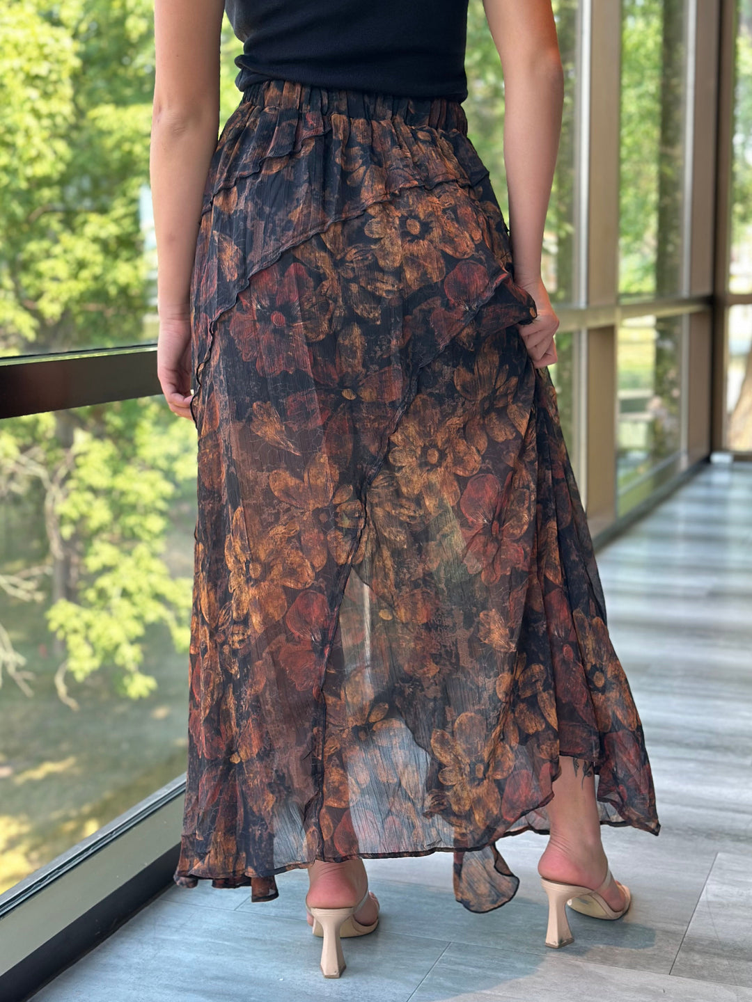 A woman wearing a long, flowy floral maxi skirt in dark brown, rust, and black tones, paired with a black top and beige heeled sandals, standing on a balcony with large windows and green trees outside.