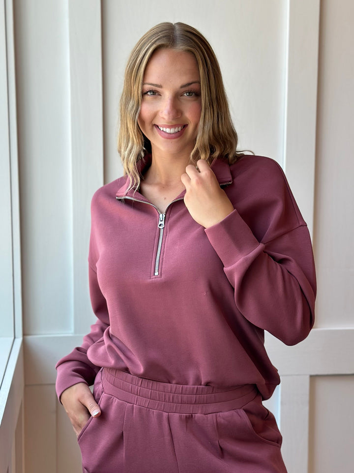 A smiling woman wearing a matching dusty rose half-zip sweatshirt and sweatpants set, standing indoors against a white paneled wall.