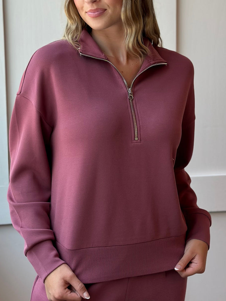 A woman wearing a dusty rose quarter-zip sweatshirt with matching sweatpants, shown from the chin down in a clean indoor setting.