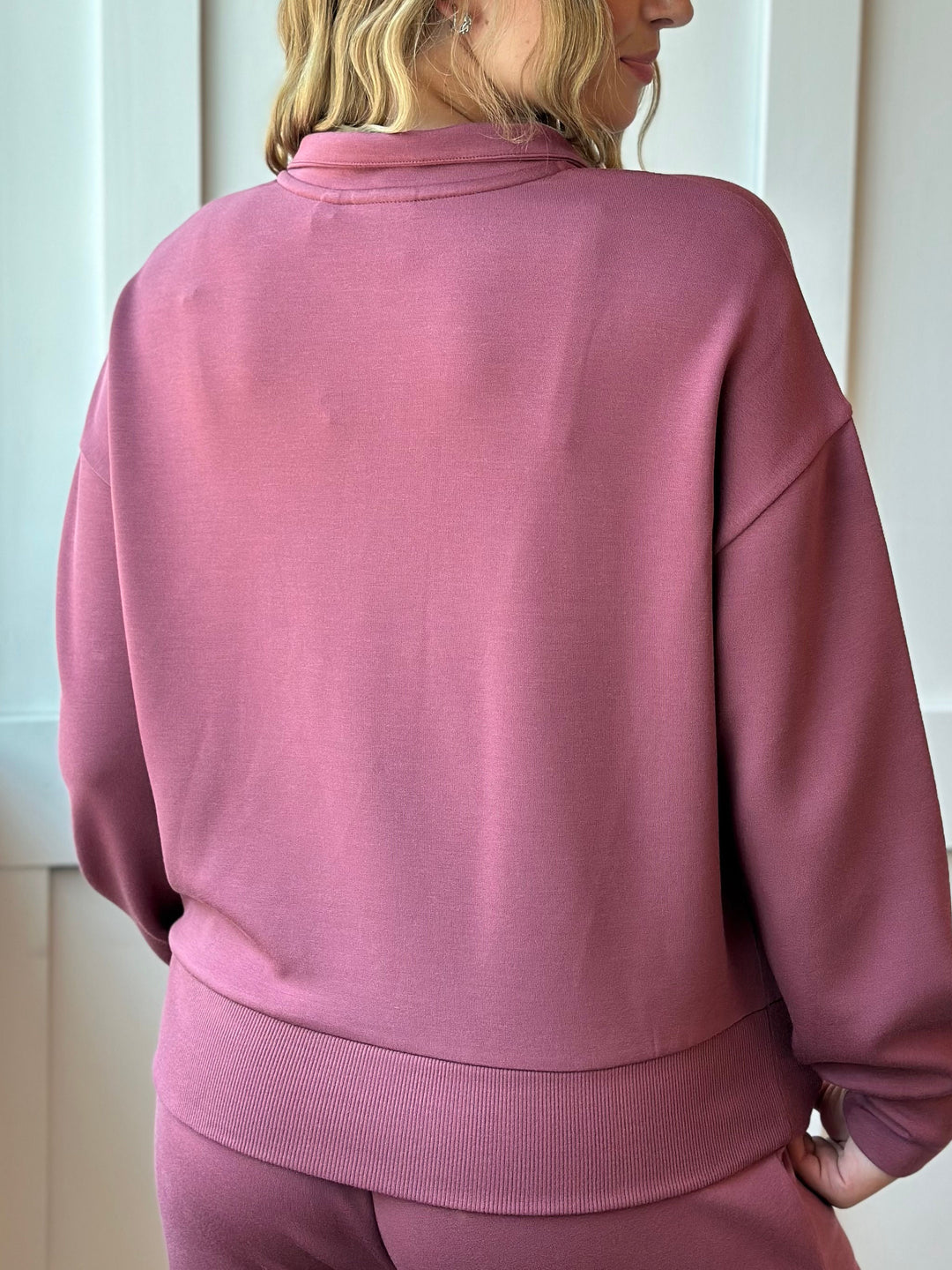 A woman shown from the back wearing a dusty rose pink sweatshirt and matching sweatpants, photographed indoors against a light neutral wall.