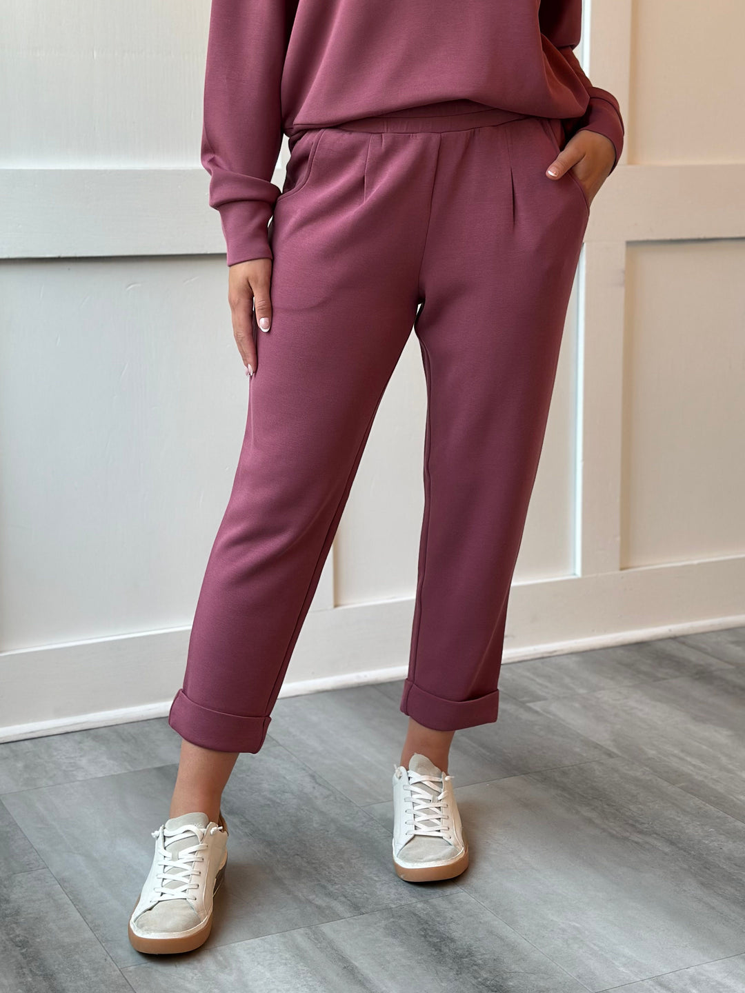 A person wearing dusty rose cropped pants with rolled cuffs, paired with a matching sweatshirt and white sneakers, standing on a gray wood floor against a white paneled wall.