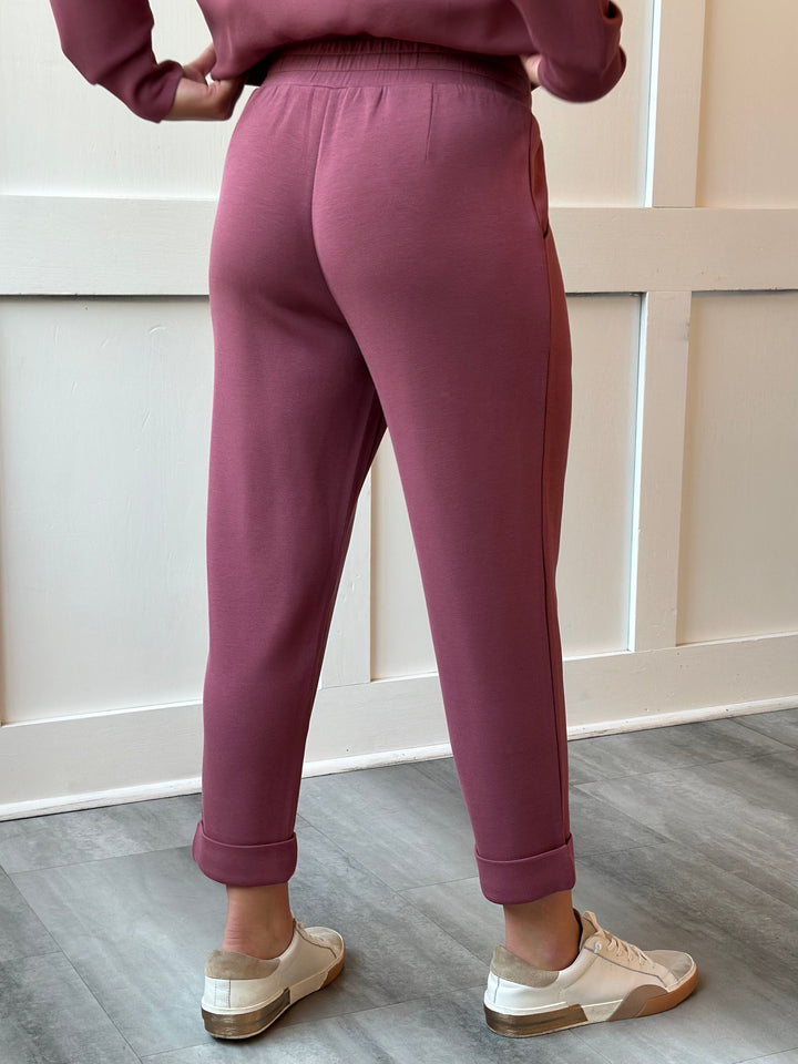 A person shown from the back wearing dusty rose jogger sweatpants with rolled cuffs, paired with a matching sweatshirt and beige sneakers, standing on a gray wood floor against a white paneled wall.