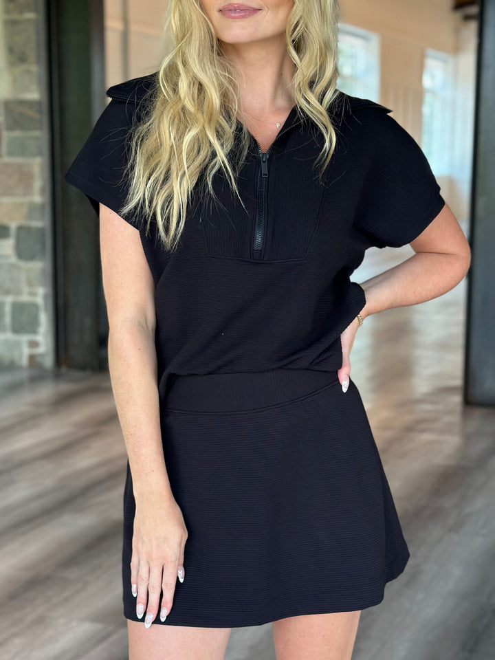 A woman wearing a black short-sleeve mini dress with a half-zip collar and a flared skirt, standing in a modern indoor space with wood floors and glass doors.