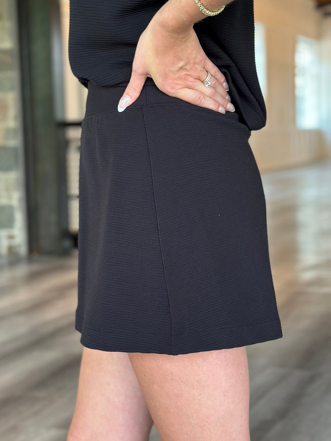 A close-up of a person wearing a black textured mini skirt with a matching black ribbed top, standing in a modern indoor hallway.